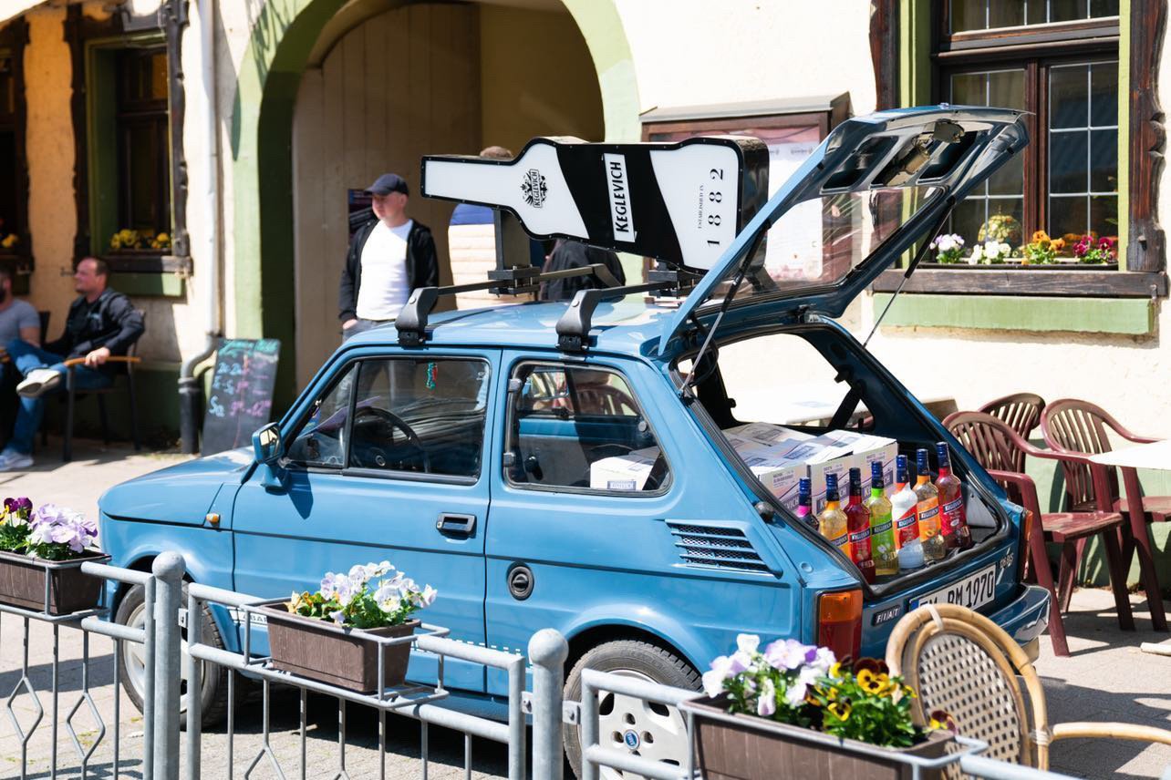 Eine entspannte Außenszene zeigt das Keglevich-Auto, ein Fiat 126 bis, in einem lebhaften blauen Farbton, das vor einem charmanten Restaurant geparkt ist. Auf dem Dach des Autos ist eine riesige Keglevich-Flasche mit Dachträgern befestigt. Der Kofferraum des Autos ist offen und enthüllt sieben Flaschen Keglevich Likör. Eine Gruppe von Menschen sitzt an einem Tisch vor dem Restaurant, teilweise sichtbar in der linken unteren Ecke. Ein gelb-schwarzes Schild, verziert mit einem Bild einer Flasche, steht vor einem Gebäude in der Mitte des Bildes. Ein Mann steht in der Nähe der linken Seite des blauen Autos und scheint mit einem Freund außerhalb des Bildes zu sprechen. Ein weißes Kreidetafel hängt an der Wand hinter dem Auto. In der rechten unteren Ecke sitzt eine Frau auf einem Rattanstuhl und bewundert eine große Topfpflanze. Das gleiche blaue Auto, mit seinem markanten weißen Rennstreifen, erscheint wieder im oberen linken Teil des Fotos. Eine Frau sitzt im Auto und schaut geradeaus. Hinter dem Auto im oberen Teil des Bildes steht eine kleine Topfpflanze. Der Blumentopf steht auf einem Metallhalter, der an einer Fensterbank des Restaurants befestigt ist. Dieses entspannte, lässige Bild lädt den Betrachter ein, den sorglosen Geist eines warmen Sommertages in einer entspannten, freundlichen Atmosphäre zu erleben.