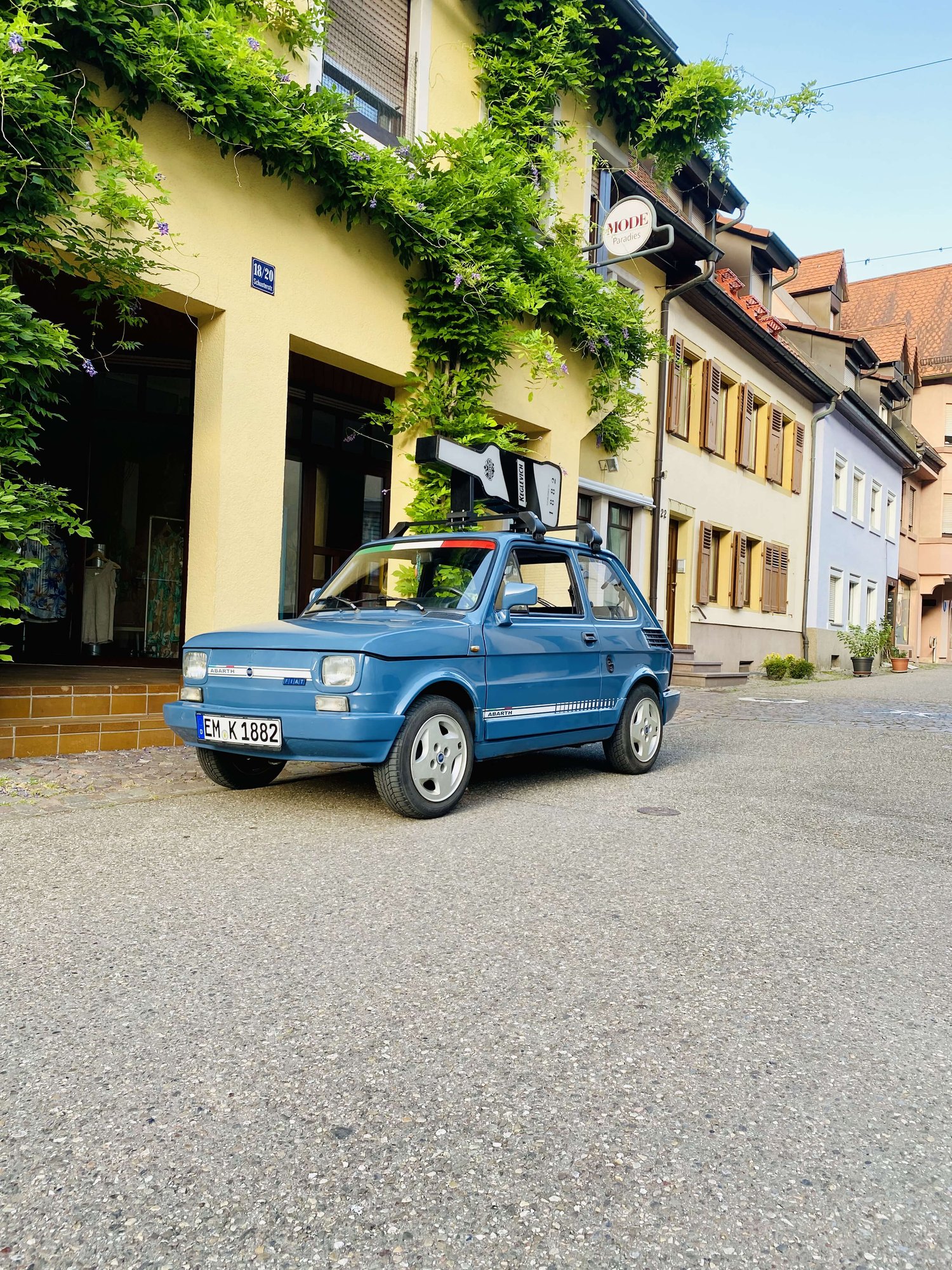 Dieses Bild zeigt eine charmante und rustikale blaue Fiat 126 bis, das Keglevich-Auto, geparkt auf der Straße neben einem malerischen Gebäude. Auf dem Dach des Autos ist ein charismatischer Dachträger befestigt, auf dem eine riesige Keglevich-Flasche platziert ist. In der Nähe des Autos fügt eine Pflanze in einem entzückenden Tontopf einen Hauch von Grün zur Szene hinzu, sie ruht auf einem rauen und unebenen Kopfsteinpflaster. Das Fahrzeug ein Gefühl von Abenteuer und Leichtigkeit aus. Insgesamt fängt das Bild die charmante Einfachheit einer malerischen Straßenszene ein und weckt ein Gefühl von Nostalgie, Sommer und Frieden.