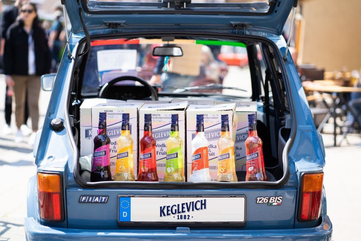 Dieses Foto zeigt den Kofferraum des lebhaft blauen Keglevich-Autos, ein Fiat 126 bis, gefüllt mit mehreren Flaschen Keglevich Likör. Rechts im Bild steht eine Gruppe von Menschen vor dem Auto und blickt zur Front des Autos.  Die Beleuchtung des Fotos ist hell und die Farben sind bunt, was ihm ein fröhliches Gefühl verleiht.